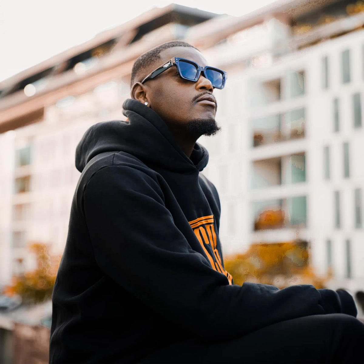 Man in a black Colonel Bench hoodie with orange CB crest and CB Shades square sunglasses
