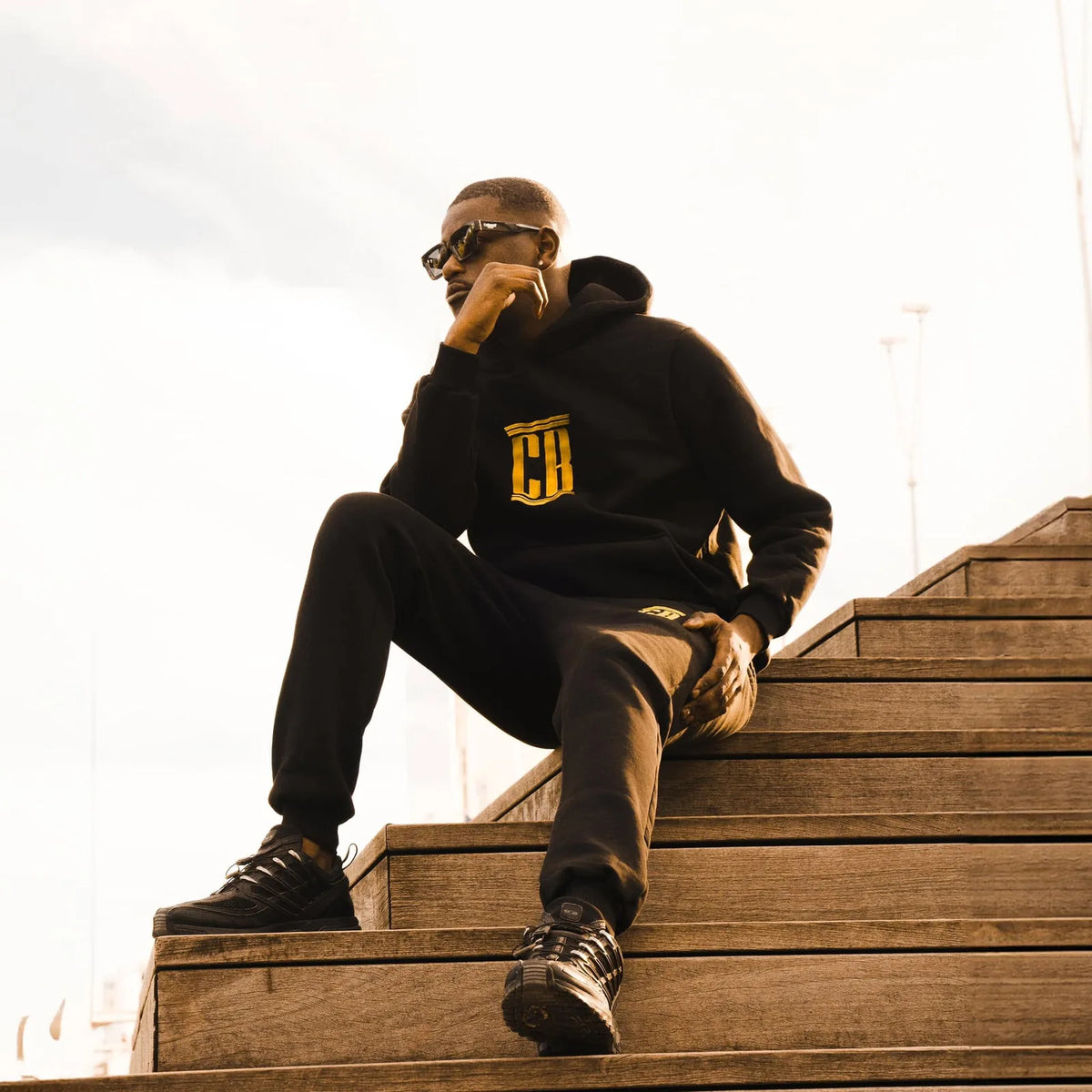 Man sitting on wooden steps in a black Colonel Bench tracksuit with gold CB logo and sunglasses