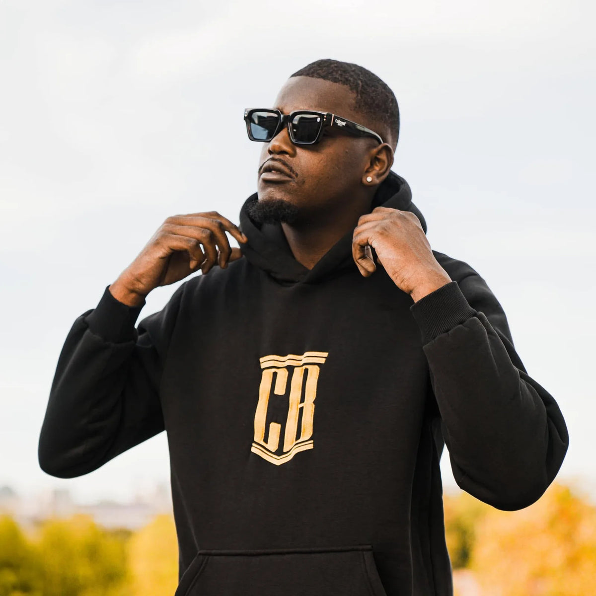 Man adjusting the hood of a black hoodie with gold embroidered CB crest, wearing CB Shades sunglasses