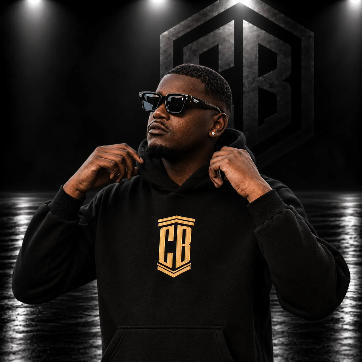 Man in a black hoodie with gold CB crest and square sunglasses, CB logo studio background