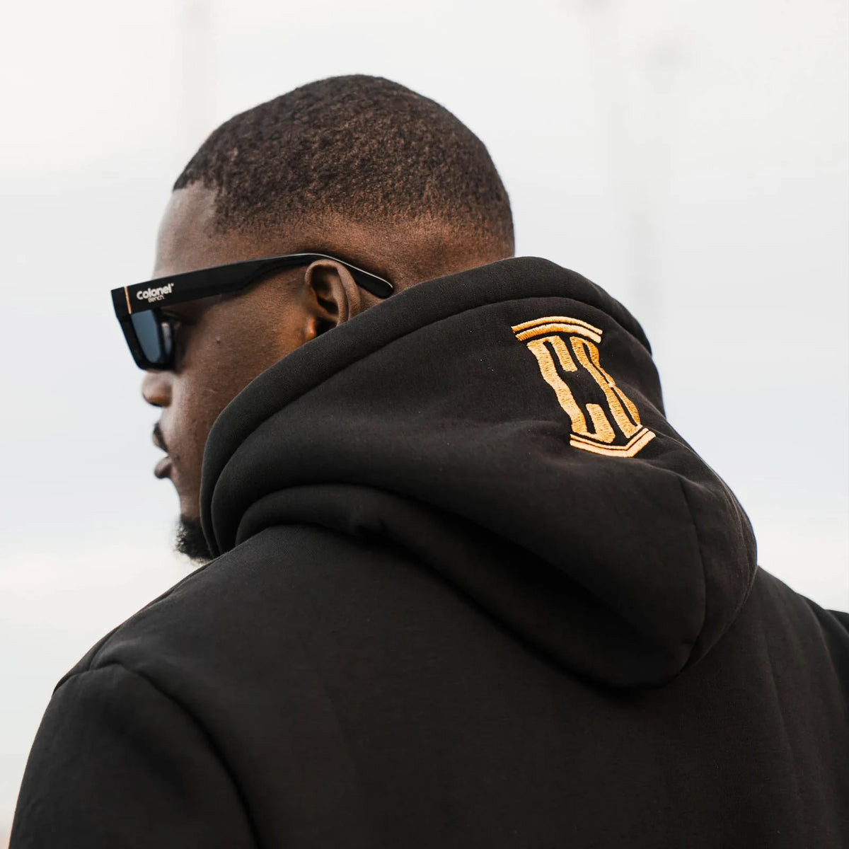 Back view of a black hoodie with gold CB embroidery on the hood, Colonel Bench sunglasses