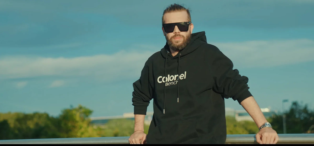 Man in a black Colonel Bench hoodie and CB Mask sunglasses leaning on a railing