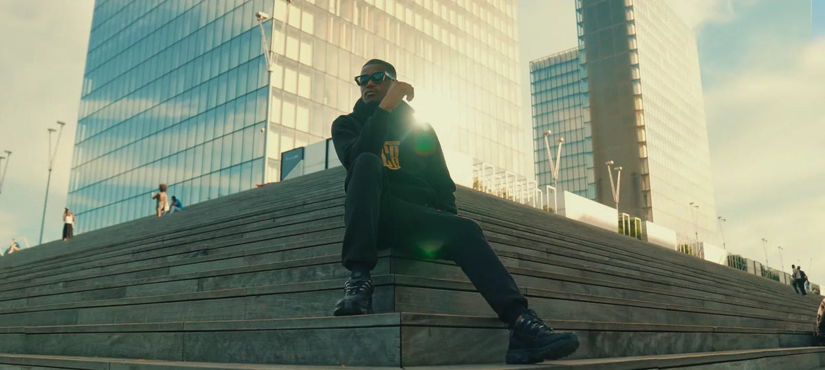 Man in a black Colonel Bench tracksuit with gold CB crest sitting on steps in the sun