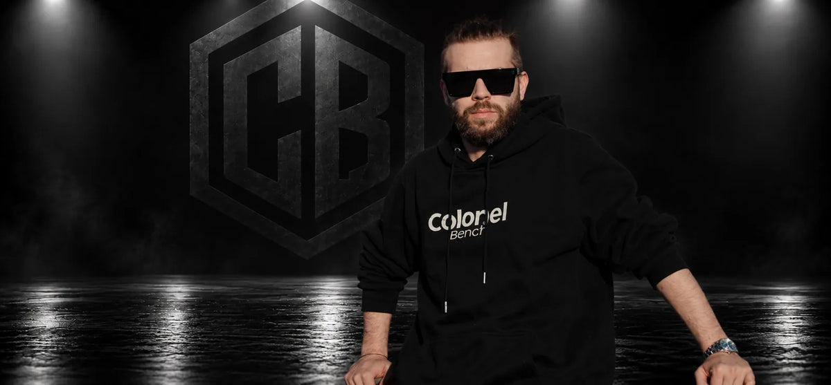 Man in a Colonel Bench wordmark hoodie and CB Mask sunglasses in front of the CB logo