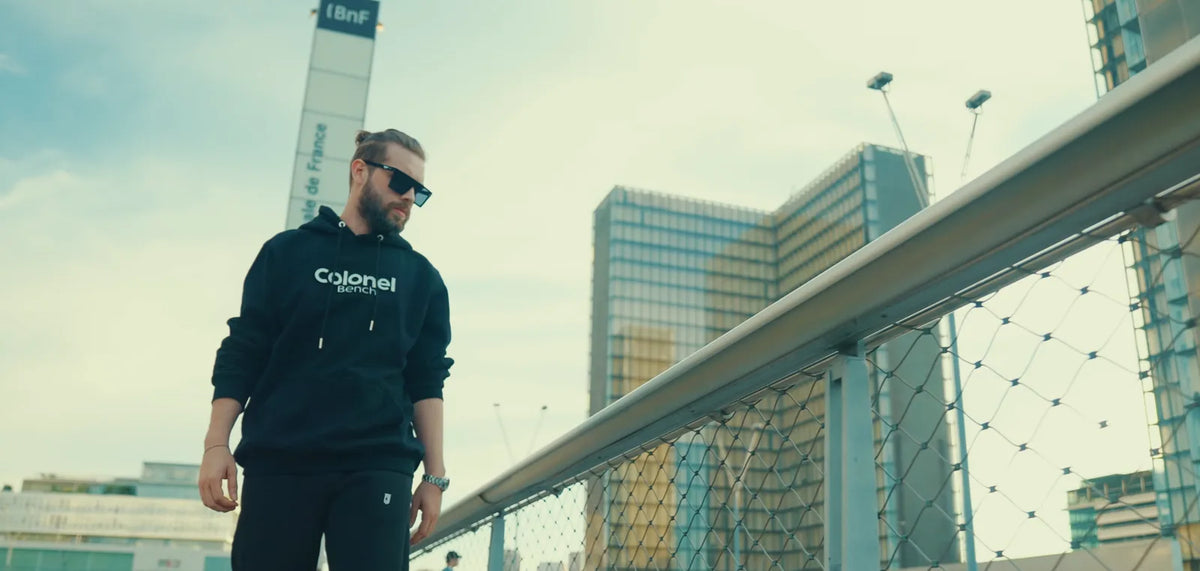 Man walking in a black Colonel Bench wordmark hoodie and CB Mask shield sunglasses
