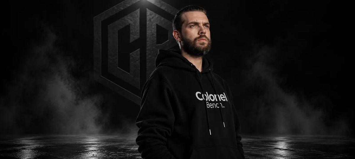 Man in a black Colonel Bench wordmark hoodie in front of the CB logo